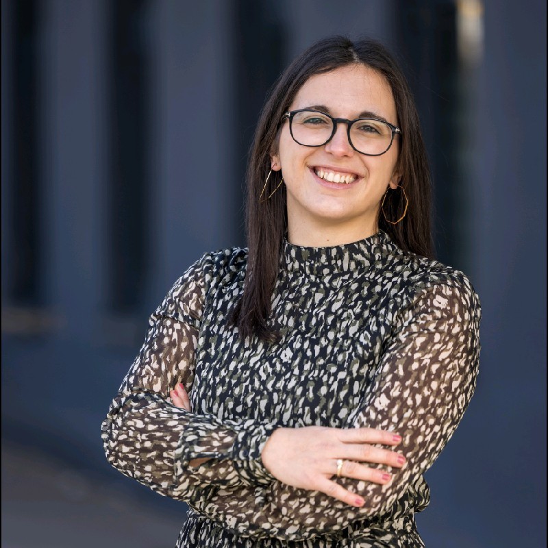 Katharina Barrett lächelt im modernen urbanen Umfeld in stilvollem Kleid und großen Brillen.